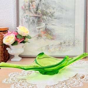 Könnte beinhalten: Eine leuchtend lindgrüne Glasschale mit einer länglichen, organischen Freiform und glatten, durchscheinenden Oberflächen. Die Schale zeichnet sich durch geschwungene, spitz zulaufende Enden aus, die nach oben ragen und eine fließende Form bilden.