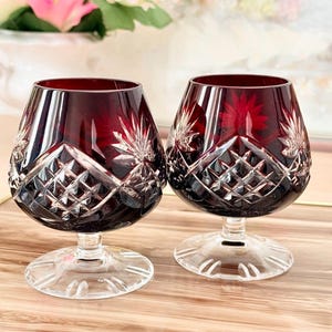 Può includere: Due bicchieri da brandy in cristallo rosso rubino con intricati disegni in vetro tagliato. Le coppe sono di un rosso intenso, che sfumano in gambi e basi trasparenti. Il vetro tagliato presenta motivi a diamante e a stella, creando un effetto scintillante. I bicchieri sono su un vassoio di legno.