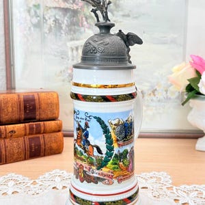 Op de afbeelding: Een vintage keramische bierpul met een gedetailleerd deksel met een soldaat te paard. De pul heeft een witte body met kleurrijke illustraties van soldaten en historische scènes, geaccentueerd met gouden randen en de tekst "in das Kaisermanöver 1908".