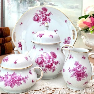 Puede incluir: Un juego de té de porcelana blanca con un vibrante patrón floral magenta y bordes dorados. El conjunto incluye una tetera con tapa, un azucarero con tapa, una jarra de leche y una bandeja grande con asas, decorados con motivos florales.