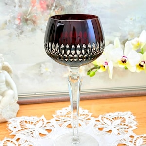 Peut inclure: Un verre à vin rouge rubis avec un pied et une base en cristal taillé transparents. Le buvant présente un motif taillé. Le verre est posé sur un napperon en dentelle blanche. Le fond comprend une peinture et des orchidées blanches.