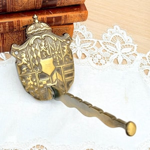Op de afbeelding: Antieke messing flesopener met een decoratief schildontwerp, met een kroon en gedetailleerde reliëfdetails. De opener heeft een lange, gebogen handgreep en een afgerond uiteinde, wat wijst op een historisch thema.