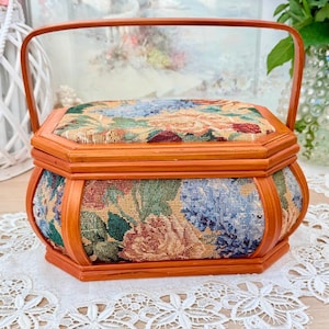 Könnte beinhalten: Eine Vintage-Nähkiste aus Holz mit Griff. Die Kiste ist achteckig mit einem floralen Wandteppichdesign an den Seiten und oben. Das Holz hat eine warme, rötlich-braune Farbe. Die Kiste steht auf einer weißen Spitzendecke.