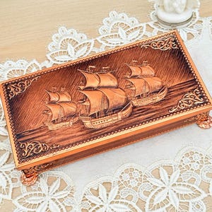 Op de afbeelding: Een rechthoekige koperkleurige doos met een gedetailleerd reliëf van drie zeilschepen op het deksel. De doos heeft sierlijke hoekontwerpen en staat op kleine, decoratieve voetjes. De achtergrond is een wit kanten kleedje.