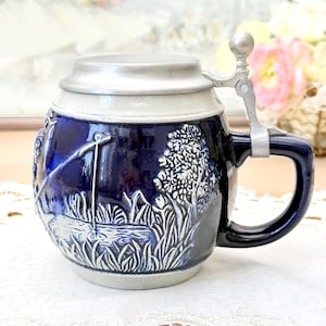 Peut inclure: Une chope à bière en céramique bleu cobalt avec une scène en relief détaillée d'une scène de pêche. La chope a un couvercle et une poignée en métal articulés. Le design comprend une scène de pêche avec un arbre et de l'eau.