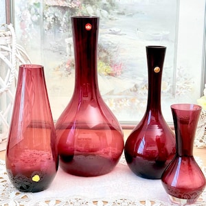 Peut inclure: Quatre vases en verre rouge rubis de différentes tailles. Les vases ont une base arrondie et un col fin. Le plus petit vase a un motif décoratif gravé à la base. Les vases sont exposés sur une nappe en dentelle blanche.