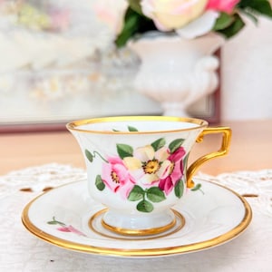 Könnte beinhalten: Ein weißes Tee-Set mit einem Blumendesign in Rosa, Gelb und Grün. Die Tasse und Untertasse haben goldfarbene Ränder. Die Tasse steht auf einem kleinen Sockel. Im Hintergrund eine Vase mit rosa und gelben Blumen.