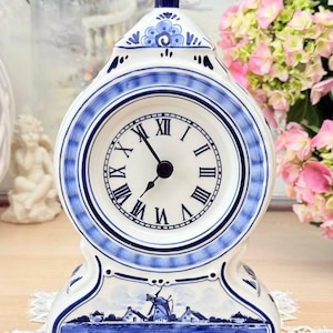Puede incluir: Un reloj de chimenea de cerámica blanca y azul con un diseño de Delftware. La esfera del reloj tiene números romanos y manecillas negras. La base presenta un molino de viento y una escena de pueblo. El reloj mide aproximadamente 20 cm de alto.