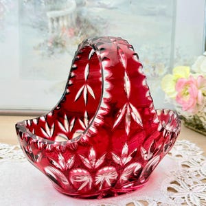 Puede incluir: Una cesta de cristal rojo rubí con asa. La cesta presenta un diseño de cristal tallado con un patrón floral. La cesta está sobre un tapete de encaje blanco. El fondo incluye una pintura y flores.