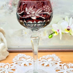 Op de afbeelding: Een robijnrood kristallen wijnglas met ingewikkelde bloemen- en druivenontwerpen in de kelk geëtst. De steel is helder en facetgeslepen, en het glas staat op een heldere, cirkelvormige voet. Het glas staat op een wit kanten kleedje. Ongeveer 20 cm hoog.