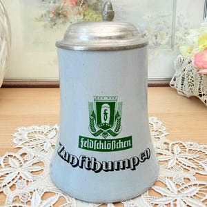 Op de afbeelding: Een lichtblauwe keramische bierpul met een metalen deksel en een decoratieve knop. De pul heeft een groen embleem met de woorden "Feldschlößchen" en "Zunftbumper" in zwarte letters. De pul staat op een wit kanten kleedje.
