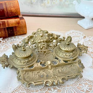 Pode incluir: Um conjunto de tinteiro de secretária em latão com dois recipientes circulares para tinta com tampa, uma escultura central em relevo de um rosto com volutas ornamentais e um suporte retangular para canetas na parte frontal.