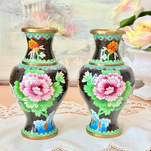 May include: A pair of matching cloisonné vases featuring a black background with intricate wirework patterns. Each vase displays vibrant pink peony flowers, green leaves, and blue rock motifs, finished with a polished brass rim and base.