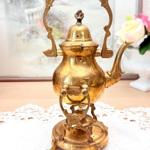 May include: A vintage brass teapot with a decorative handle and spout. The teapot sits on a matching stand with ornate detailing. The gold-colored metal has a polished finish, reflecting light. The teapot is likely for decorative purposes.