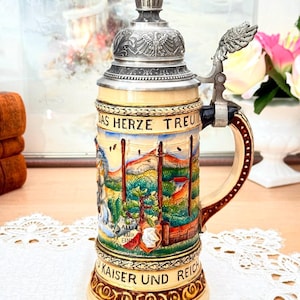 Peut inclure: Une chope à bière allemande vintage avec un couvercle et une poignée argentés. Le corps en céramique présente une scène de paysage peinte avec le texte "Das Herze Treu" et "Kaiser Und Reich". La chope a une poignée décorative et un design détaillé.