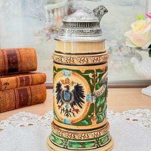 Può includere: Un boccale da birra decorativo con coperchio metallico incernierato. Il corpo in ceramica presenta un design dettagliato con uno stemma, un'aquila e dettagli verdi e dorati. In stile tradizionale tedesco.