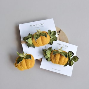 Könnte beinhalten: Handgemachte Haaraccessoires in Form von Kürbissen aus Filz mit grünen Blättern und Stielen. Die Kürbisse sind leuchtend orangefarben. Die Accessoires werden auf weißen Karten mit dem Text "MAKATUSIA Felt Flowers Handmade Accessories" präsentiert.
