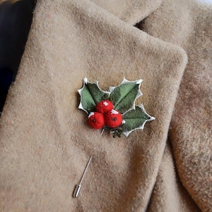 May include: A festive holly brooch with three red berry clusters and green leaves with white accents. The brooch is pinned to a tan wool coat. A silver pin is attached to the brooch.