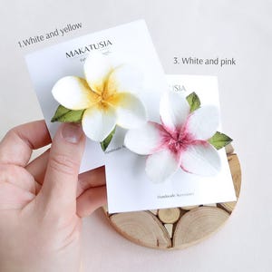 May include: Two handmade plumeria flower hair clips. One is white and yellow, the other is white and pink. Each flower has green leaves and is attached to a white card with the brand name "MAKATUSIA" printed on it.