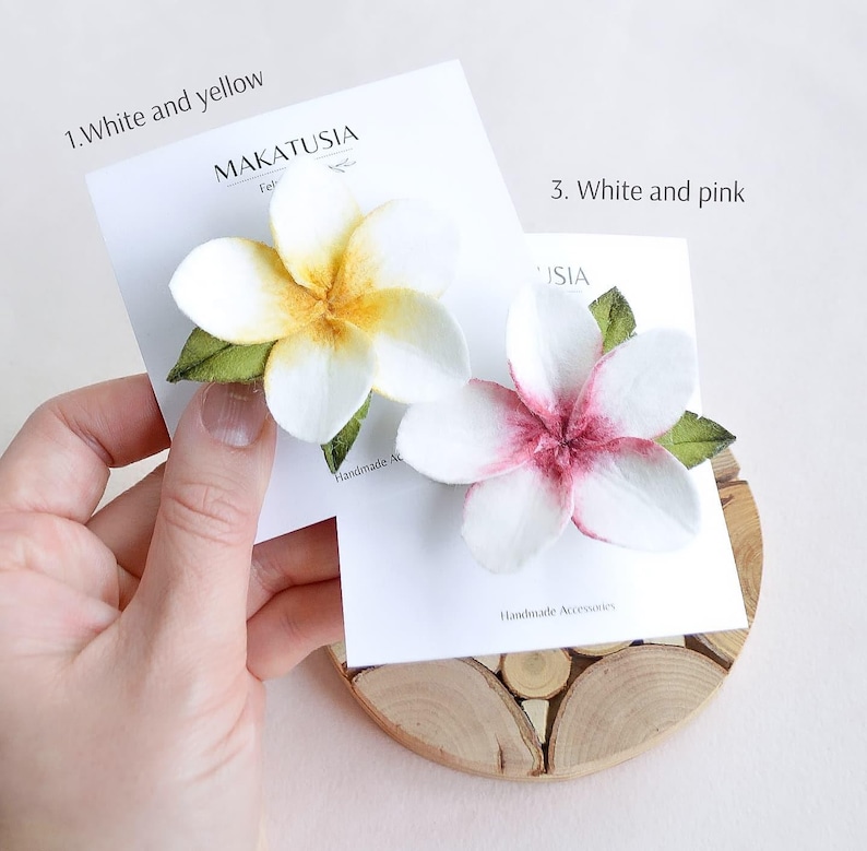May include: Two handmade plumeria flower hair clips. One is white and yellow, the other is white and pink. Each flower has green leaves and is attached to a white card with the brand name "MAKATUSIA" printed on it.