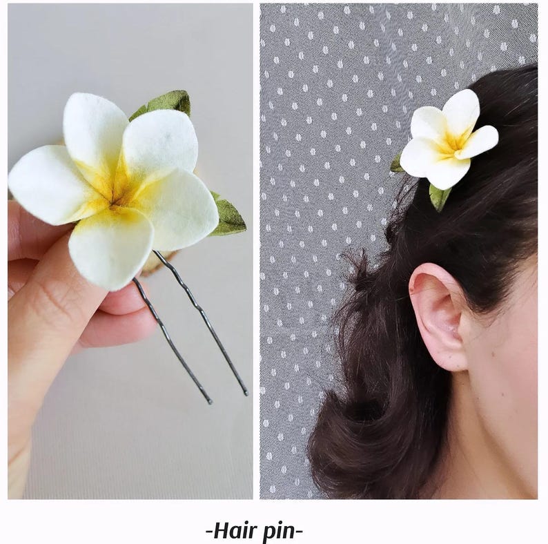 May include: A white and yellow plumeria flower hair pin with green leaves. The hair accessory is shown held by a hand and worn in dark hair. The text "-Hair pin-" is at the bottom of the image.