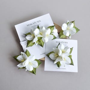 May include: White felt flower hair accessories with green leaves, displayed on white cards. The cards have the text "MAKATUSIA Felt Flowers Handmade Accessories". The flowers are clustered together, creating a natural, floral design.