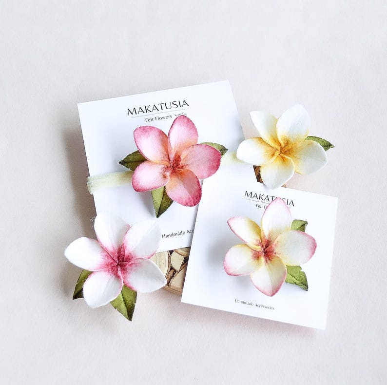 May include: Four felt flower hair accessories in pink, white, and yellow with green leaves. Each flower is attached to a white card with the text "MAKATUSIA Felt Flowers" and "Handmade Accessories". The flowers are arranged on a light background.