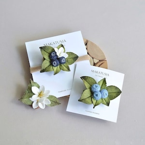 May include: Handmade felt flower hair accessories. The image shows three hair accessories with a blueberry and flower theme. The accessories are attached to white cards with the text "MAKATUSIA Felt Flowers Handmade Accessories". The flowers are white, blue, and green.