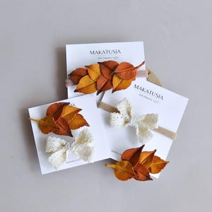 Pode incluir: Três tiaras com desenhos de folhas e laços de feltro. As folhas são em tons de laranja e amarelo, enquanto os laços são brancos. Cada tiara é exibida em um cartão branco com o texto "MAKATUSIA Felt Flowers".
