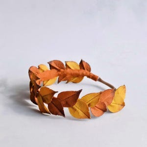 May include: A handmade leaf crown featuring a circular design with brown, yellow, and orange leaves. The leaves are arranged around a brown stem, creating a natural and rustic aesthetic. Perfect for fall-themed events or photo shoots.