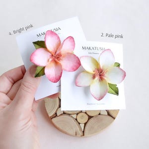 May include: Two felt flower hair accessories, one bright pink and one pale pink, each with green leaves. The flowers are displayed on white cards with the text "MAKATUSIA Felt Flowers Handmade Accessories". The accessories rest on a wooden coaster.