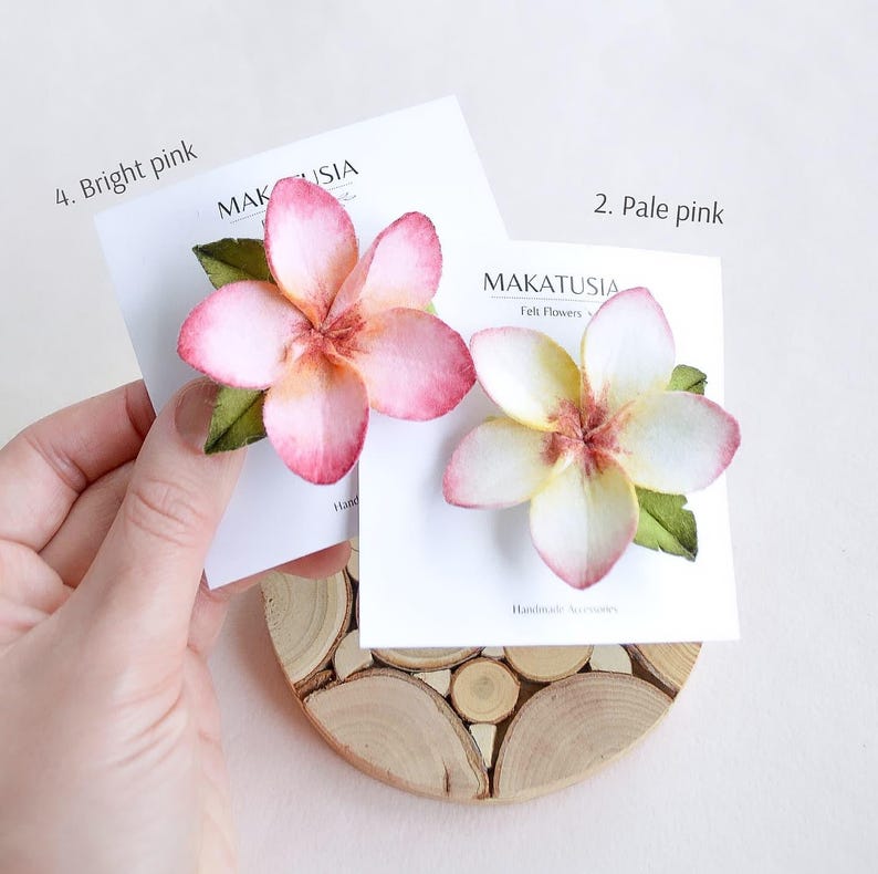 May include: Two felt flower hair accessories, one bright pink and one pale pink, each with green leaves. The flowers are displayed on white cards with the text "MAKATUSIA Felt Flowers Handmade Accessories". The accessories rest on a wooden coaster.