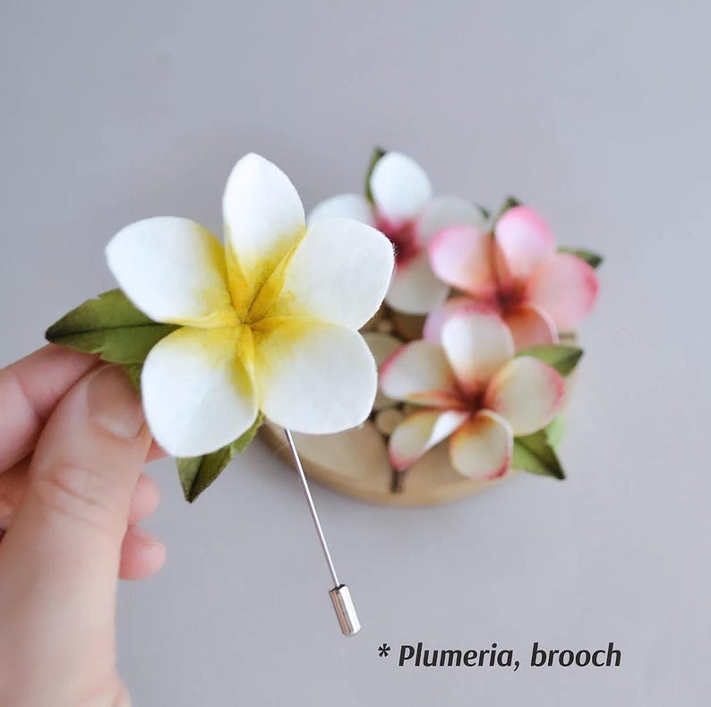 May include: A handmade plumeria brooch featuring white and pink fabric flowers with yellow centers and green leaves. The brooch has a silver pin and the text "* Plumeria, brooch".