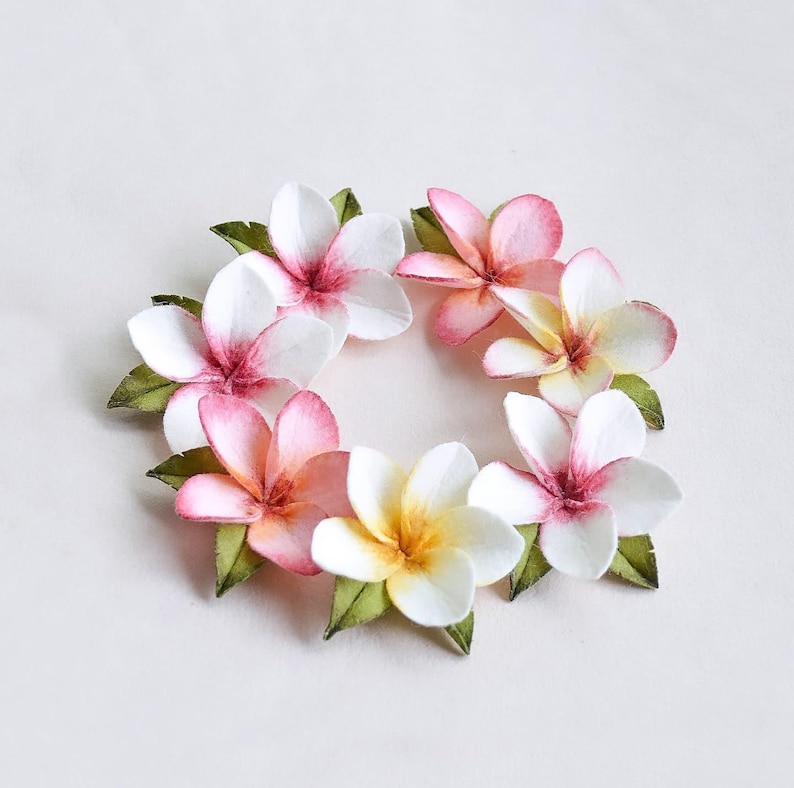 May include: A circular arrangement of paper flowers. The flowers are white, pink, and yellow with green leaves. The flowers have a realistic appearance and are arranged in a wreath-like pattern on a white surface.