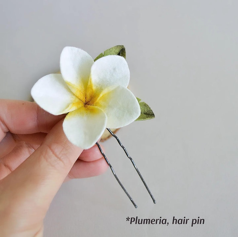 May include: A white plumeria flower hair pin with a yellow center and green leaves. The hair accessory is held by a hand. The text "*Plumeria, hair pin" is at the bottom of the image.