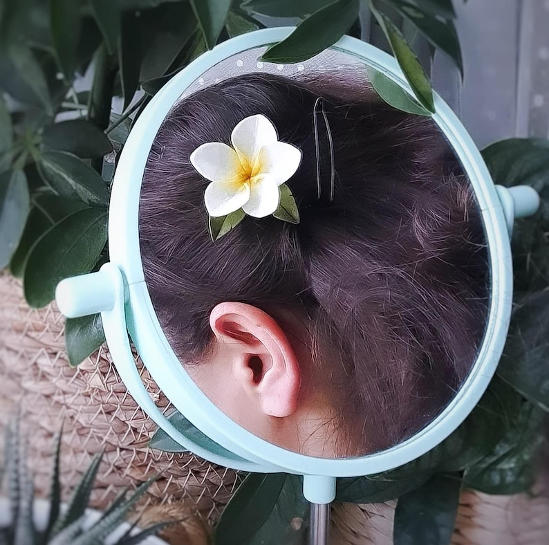 May include: A light blue, round, handheld mirror reflects a person's hair, ear, and a white and yellow flower hair accessory. The mirror is surrounded by green leaves and foliage, creating a natural, botanical theme.
