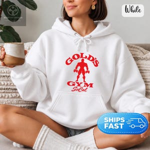 Könnte beinhalten: Ein weißer Kapuzenpullover mit einem roten Grafikdruck auf der Brust. Das Motiv zeigt die Silhouette eines Bodybuilders mit Hanteln, wobei der Text Gold's Gym SoCal in einer fetten roten Schrift über und unter der Figur steht.
