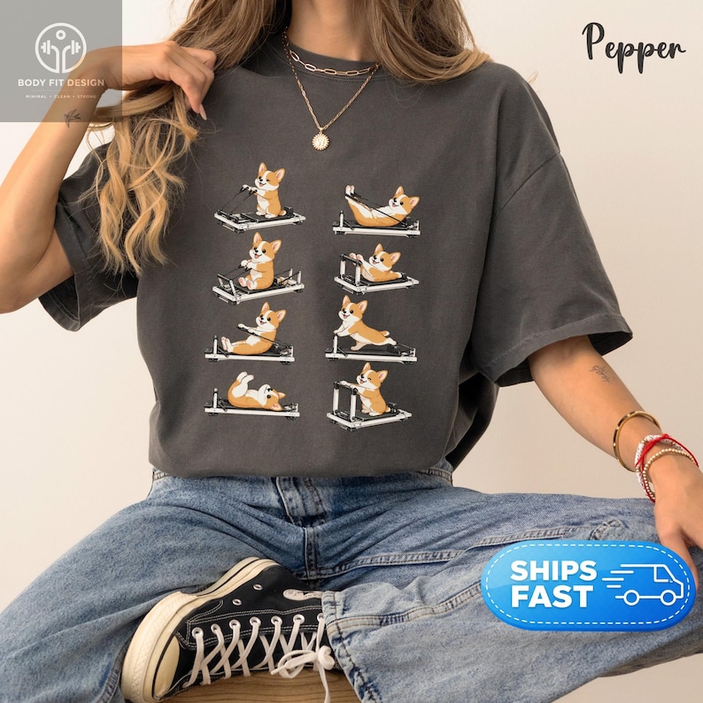 Op de afbeelding: Donkergrijs T-shirt met een ontwerp van corgi-honden die oefeningen doen op Pilates-reformer. De corgi's worden in verschillende poses getoond, met het woord "Pepper" in script bovenaan rechts. Het shirt heeft een losse pasvorm.