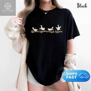 Peut inclure: T-shirt noir avec un motif humoristique de quatre oies blanches sur des tapis roulants. Les oies sont repr&eacute;sent&eacute;es dans diff&eacute;rentes poses, ajoutant une touche d'humour. Le t-shirt a un col rond et des manches courtes.