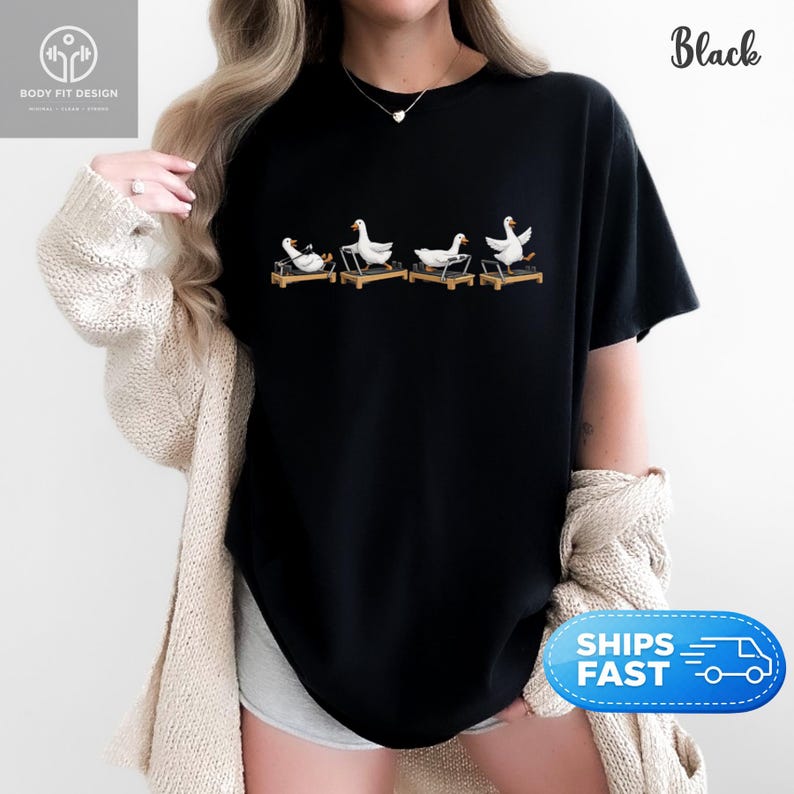 Peut inclure: T-shirt noir avec un motif humoristique de quatre oies blanches sur des tapis roulants. Les oies sont repr&eacute;sent&eacute;es dans diff&eacute;rentes poses, ajoutant une touche d'humour. Le t-shirt a un col rond et des manches courtes.
