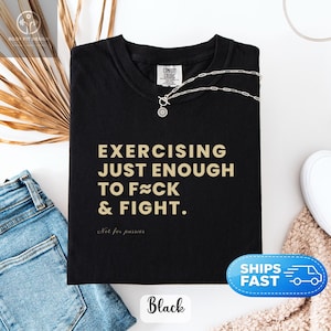 Può includere: Una t-shirt nera a maniche corte con il testo dorato EXERCISING JUST ENOUGH TO F≈CK & FIGHT. e il testo più piccolo in corsivo Not for pussies. La maglietta presenta un girocollo e un'etichetta del marchio Comfort Colors sul colletto interno.