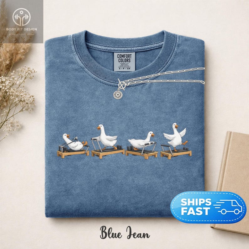 Peut inclure: T-shirt bleu jean &agrave; col ras du cou. Le t-shirt pr&eacute;sente un motif de quatre oies blanches faisant de l'exercice sur des appareils de Pilates. Le texte "Blue Jean" est en bas. Un collier argent&eacute; est sur le t-shirt.