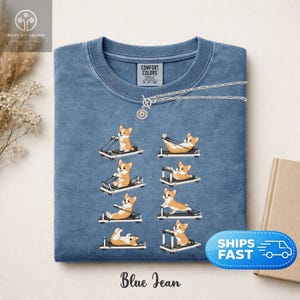 Op de afbeelding: Blauw spijkerbroekkleurig T-shirt met een ronde hals. Het shirt heeft een afbeelding van corgi-honden die oefeningen doen op pilatesmachines. De tekst "Blue Jean" staat onderaan het shirt. Een zilveren ketting is over de bovenkant van het shirt gedrapeerd.