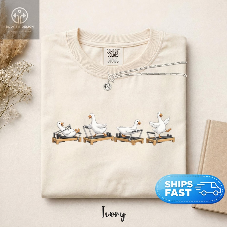 Peut inclure: T-shirt ivoire avec un motif fantaisiste de oies blanches faisant de l'exercice sur des appareils de Pilates. Le t-shirt a un col rond et des manches courtes. Un collier argent&eacute; avec un pendentif soleil est drap&eacute; sur le t-shirt. Le mot "Ivory" est en bas.