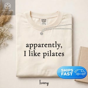 May include: Ivory-colored t-shirt with the phrase "apparently, I like pilates" in black lettering. A silver necklace with a sun pendant is draped over the shirt. The shirt is folded and displayed on a neutral surface.