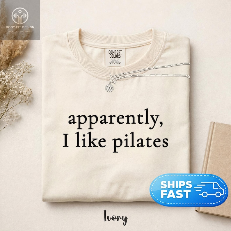 May include: Ivory-colored t-shirt with the phrase "apparently, I like pilates" in black lettering. A silver necklace with a sun pendant is draped over the shirt. The shirt is folded and displayed on a neutral surface.