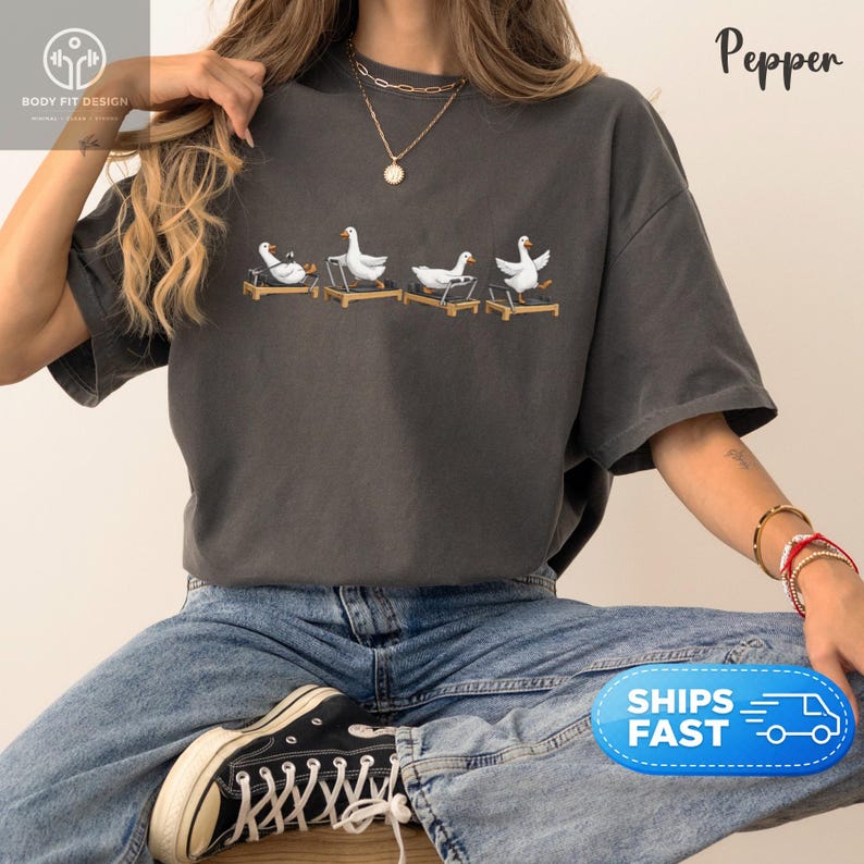 Peut inclure: T-shirt gris fonc&eacute; avec un motif brod&eacute; de quatre oies blanches sur des tapis roulants. Le mot "Pepper" est imprim&eacute; en &eacute;criture cursive. Le t-shirt est port&eacute; avec un jean bleu et des baskets noires.