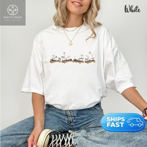 Peut inclure: T-shirt blanc avec un motif fantaisiste de oies faisant de l'exercice sur des tapis roulants. Le dessin est de style cartoon, avec les oies dans diverses poses. Le t-shirt a une coupe d&eacute;contract&eacute;e et est fait d'une mati&egrave;re douce.