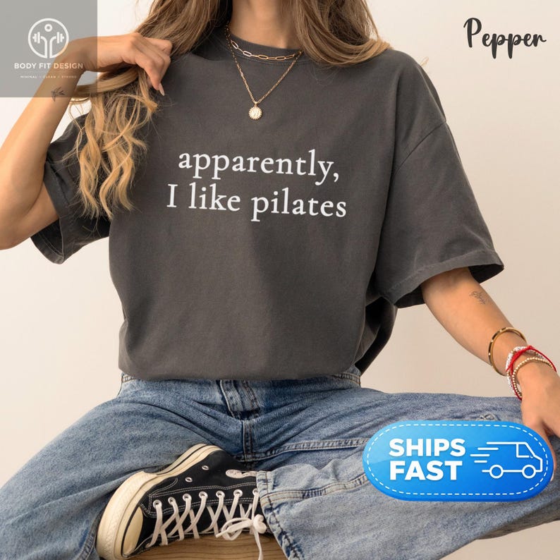 May include: Dark gray t-shirt with the text "apparently, I like pilates" in white. The shirt is paired with blue jeans and black sneakers. The image features a casual, relaxed style, suitable for fitness enthusiasts.