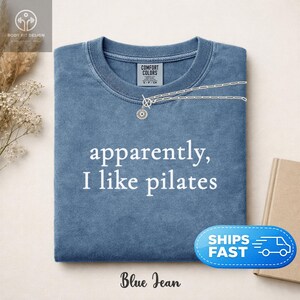 May include: Blue jean colored t-shirt with the text "apparently, I like pilates" in white. A silver necklace with a sun pendant is draped across the shirt. The shirt has a crew neck and is folded. The words "Blue Jean" are at the bottom.